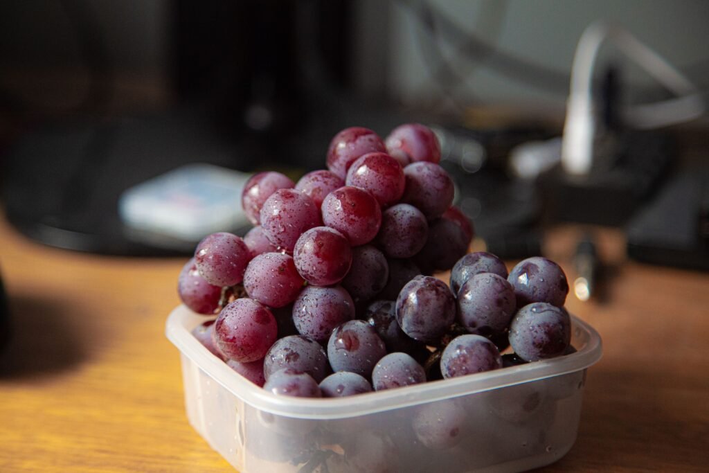 Cacho de uvas roxas dentro de pote plástico sobre mesa de madeira