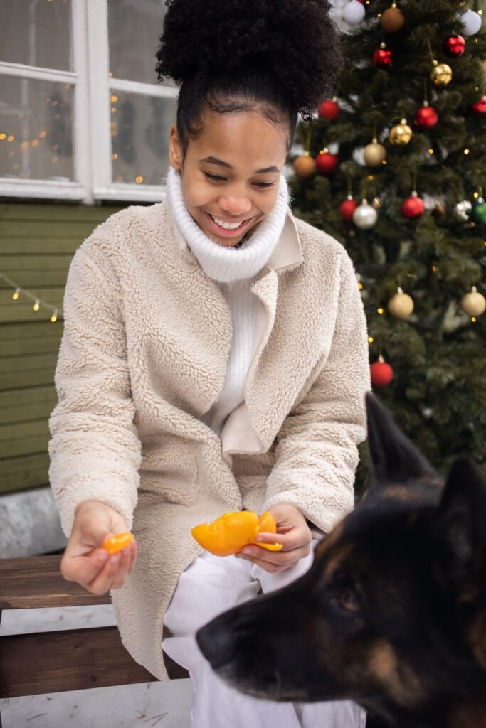 Mulher sorridente segurando gomos de tangerina descascada na mão, com a cabeça de um cachorro em primeiro plano e uma árvore de Natal decorada ao fundo