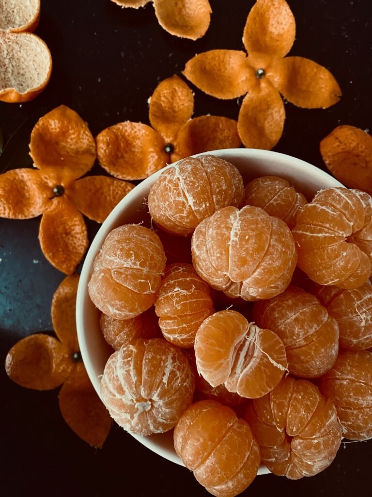 Tigela branca cheia de tangerinas descascadas, cercada por cascas de tangerina dispostas em formato de flor sobre uma superfície escura